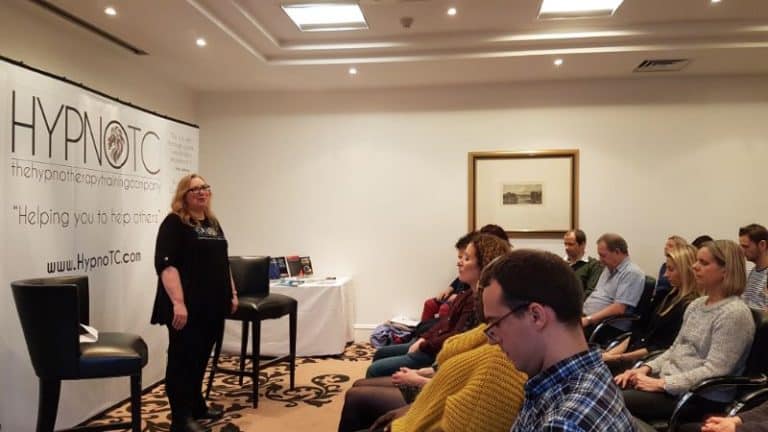 Kate talking to a group, by the HypnoTC banner - Hypnotherapy training for healthcare professionals